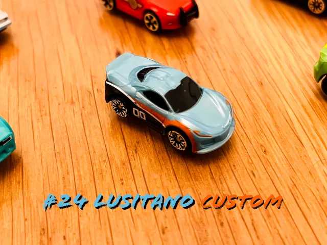 A carousel photo displaying the twenty-fourth vehicle named 'Lusitano Custom' out of an array of 'Deluxe Cars', at full size. The car is painted mostly grey with an orange streak of paint across the right side door and front wheel.